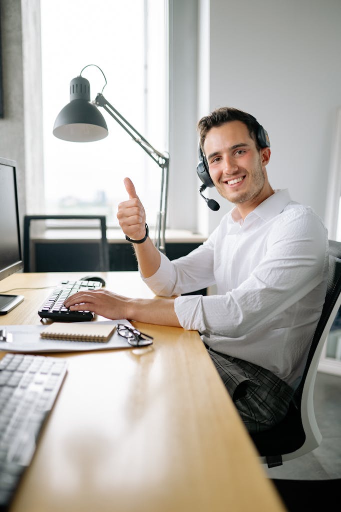 Friendly customer service agent wearing headset and smiling with a thumbs up at a modern office desk.